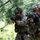 Two soldiers in full multicam combat gear are moving through a wooded area with weapons raised in a tactical formation. Both wear helmets with accessories and carry what appear to be training weapons indicated by yellow/orange safety markers. Additional personnel are visible in the background, suggesting a military training exercise.