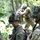 The image shows U.S. Army soldiers in a wooded training environment wearing Operational Camouflage Pattern (OCP) uniforms and full combat gear. The foreground soldier displays a 4th Infantry Division shoulder patch and is equipped with a helmet-mounted night vision device mount, ballistic eyewear, and a tactical watch. Multiple soldiers are visible conducting what appears to be a field training exercise.