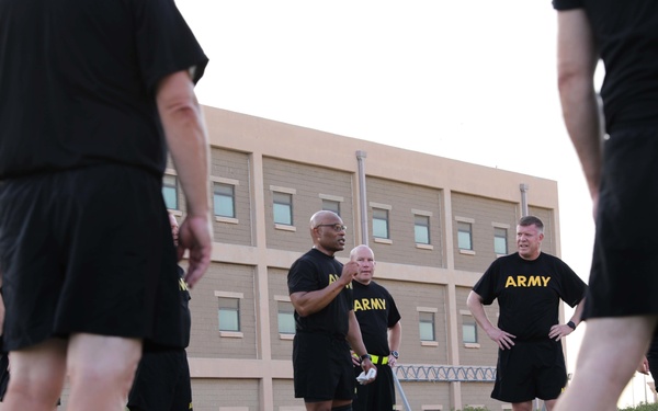 A group of U.S. Army personnel are gathered on an outdoor artificial turf field, wearing matching black PT (Physical Training) uniforms with yellow 'ARMY' lettering. The image is taken from a low angle, with several individuals in the foreground and a small group appearing to converse or receive instruction in the middle ground. A multi-story tan/brick building is visible in the background, suggesting a military installation.