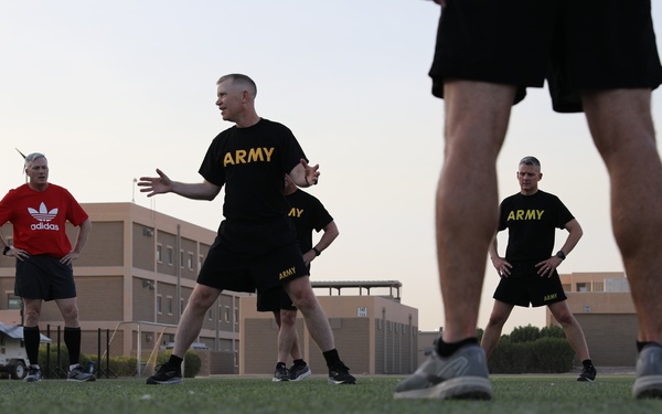 A group of U.S. Army personnel are conducting physical training (PT) on a grass field, likely at a military installation. One individual in the center appears to be leading or instructing the group, with arms extended in a demonstrative gesture. The setting appears to be early morning or dusk based on the ambient lighting.
