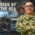 A U.S. Navy service member in digital camouflage (NWU Type III) uniform stands with arms crossed, smiling in front of a Navy recruiting poster. The poster reads 'Forged by the Sea' and displays the website 'NAVY.COM'. The individual's name tape reads 'DUTTON' and the branch tape reads 'U.S. NAVY'.