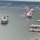An aerial view of a busy inland waterway port showing multiple large bulk carrier vessels docked or navigating the river channel. Several covered barges are moored along the right bank, and industrial port infrastructure including a bridge is visible in the background. The scene depicts active commercial maritime operations on what appears to be a major river system.