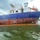 A large commercial vessel with a blue hull and white superstructure is moored at a waterway facility. The ship's hull shows significant rust and paint deterioration along the waterline, and an orange containment boom is deployed around the vessel in the water. The scene suggests an environmental response or pollution containment operation is underway.