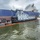 A tugboat named 'Justice' is positioned alongside a large blue-hulled cargo/bulk carrier vessel on a waterway. An orange oil containment boom is deployed around the vessels, indicating an active spill response or precautionary containment operation. A work barge with pumping equipment is also visible on the left side of the scene.