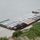 An aerial view shows a large string of loaded river barges moored along a riverbank, with cargo holds covered by tarpaulins in various colors. The barges appear to be grounded or very close to the shoreline, with some flooding or shallow water visible between the vessels and the bank. Construction or salvage equipment is visible on the shore below.
