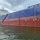 A large cargo or tanker vessel is shown moored in a body of water, displaying significant hull weathering and paint deterioration. An orange containment boom is deployed along the waterline, suggesting an active pollution containment or spill response operation. The vessel shows a blue upper hull and red anti-fouling lower hull, with visible draft markings on the starboard side.