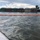 The image shows a river spill response operation with an orange containment boom deployed across the water surface near a fleet of moored barges. The barges have green-covered cargo holds with dome-shaped covers, and a control tower or crane structure is visible in the background. Dark streaks on the water surface suggest the presence of an oil or fuel spill being contained by the boom.