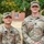 Two U.S. Army soldiers in Operational Camouflage Pattern (OCP) uniforms are posing together outdoors near a tree and brick building. The soldier on the left is identified by name tape as 'ALMARAZ' and the taller soldier on the right is identified as 'MEADE', who displays a First Lieutenant rank insignia on his cap. Both are smiling and appear to be on a military installation.