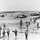 A black-and-white photograph depicting a large-scale amphibious military landing operation, with numerous soldiers wading through shallow surf toward a beach. Multiple landing craft vessels are visible in the background, having deployed troops into the water. The soldiers are heavily equipped with full combat gear, helmets, and weapons.