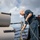 A U.S. Navy sailor in blue working uniform is hosing down equipment on the deck of a naval vessel at sea. The sailor is using a brass-nozzle hose to wash the base of what appears to be torpedo tube launchers or similar cylindrical deck-mounted weapons systems. The ocean and partly cloudy sky are visible in the background, indicating the ship is underway.