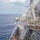 Navy sailors are conducting a freshwater washdown of a U.S. Navy warship, likely a guided-missile destroyer or cruiser, while underway at sea. Multiple personnel are visible operating orange fire hoses to wash down the exterior superstructure and deck of the vessel. A small rainbow is visible in the water spray near the center of the image.