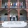 Three military personnel in uniform are standing on the steps of the Inter-American Defense College, a red brick building with arched architecture. Multiple national flags from various countries are displayed on the upper balcony of the building. The scene appears to be an official or ceremonial occasion at this multinational defense institution.