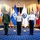 Three military officers in different uniforms stand formally in what appears to be an official ceremonial hall. They are positioned in front of a large display of international flags, including a prominently displayed Honduran flag and an organizational seal/flag. The setting suggests a multinational military or diplomatic event.
