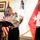 Two military officers are present in what appears to be an official signing ceremony in a formal office setting. One officer is signing a document on a lectern while the other stands at attention observing. Multiple flags are visible in the background, including a US flag and what appears to be an Inter-American Defense Board flag.