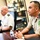 Two military officers are seated at a conference table in an office setting, appearing to be engaged in or attending a meeting. The officer in the foreground wears a tan/khaki uniform with a distinctive green and gold circular patch, while the officer in the background wears a white U.S. Army-style uniform. The setting appears to be a formal military office with display cases and awards visible in the background.