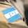 A close-up photograph of an embroidered flag patch sewn onto a tan/khaki uniform sleeve. The patch depicts the flag of Honduras, featuring two horizontal blue stripes, a white center stripe, and five blue stars arranged in an X pattern. The image is sharp in the foreground with a shallow depth of field blurring the background.