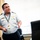 A uniformed official, likely a military or aviation officer, is standing in an office environment and appears to be speaking or presenting. He is wearing a light gray short-sleeve uniform shirt with gold epaulettes, a wing insignia badge, and an ID lanyard. The setting appears to be an administrative office with a desk, keyboard, and office chairs visible.