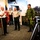 A group of military personnel from multiple nations are gathered in what appears to be a modern military briefing room or lecture hall. A bald officer in a white dress uniform with black stripe trousers appears to be speaking or briefing the group, which includes personnel in tan/khaki uniforms and one in Canadian Forces CADPAT camouflage pattern. A Colombian flag is visible in the background, suggesting an international military exchange or liaison visit.