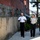 The image shows a group of military and uniformed personnel walking along a paved pathway beside a brick and stone building. Three individuals are prominently in the foreground, wearing different military uniforms suggesting a multinational or inter-agency meeting. The setting appears to be a military installation or government facility.