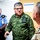 The image shows three individuals in a corridor or hallway setting, with two wearing military uniforms and one in a police or security uniform. The central figure, wearing green digital camouflage with 'EJERCITO MEXICANO' (Mexican Army) insignia, appears to be engaged in conversation with a third individual in tan/khaki military attire. A building evacuation plan is visible posted on the wall in the background.