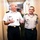 Two military officers in dress uniforms walk together down an indoor corridor, engaged in conversation. A third uniformed individual is visible in the background. The setting appears to be a military or government facility.
