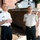 Two military officers in uniform are standing outdoors in front of a brick building, appearing to be engaged in a friendly conversation. The officer on the left wears a white US Army dress uniform with two-star general rank insignia, while the officer on the right wears a tan/khaki uniform with green trousers and red collar insignia suggesting a foreign military affiliation. The setting appears to be a formal military installation or institutional campus.