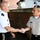 Two military officers in uniform are shaking hands outdoors in front of a brick building. One officer wears a white US Army uniform with two-star major general rank insignia, while the other wears a tan/khaki uniform with what appears to be a foreign military's insignia and a green circular shoulder patch. The interaction appears to be a formal military greeting or diplomatic exchange.