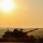 1-68 AR conducts an Abrams platoon live-fire exercise A military main battle tank is silhouetted against a hazy golden sunset or sunrise sky. At least one crew member is visible in the open turret hatch, and a small flag or pennant is mounted on the vehicle. The background features a flat, open landscape with a treeline obscured by atmospheric haze.