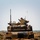 1-68 AR conducts an Abrams platoon live-fire exercise A rear view of a desert-tan painted main battle tank, consistent with an M1 Abrams, positioned on a dirt berm or earthen mound. A single soldier is visible in the commander's hatch, wearing a helmet and appearing to operate or observe from the turret. The setting appears to be a training range or field exercise environment based on the terrain and surrounding landscape.