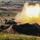 1-68 AR conducts an Abrams platoon live-fire exercise A main battle tank is firing its main gun from a hull-down defensive position behind an earthen berm. A large muzzle blast with bright orange and yellow flames and white smoke is visible at the end of the barrel. The setting appears to be a military training range or exercise in an open, semi-arid landscape.