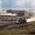 1-68 AR conducts an Abrams platoon live-fire exercise The image shows multiple main battle tanks maneuvering across an open, dusty terrain during what appears to be a military exercise or training operation. At least three tanks are visible moving in formation, generating significant dust clouds as they traverse the dry, scrubby landscape. The scene is consistent with a large-scale armored training exercise conducted in an open field environment.