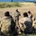 1-68 AR conducts an Abrams platoon live-fire exercise A military instructor or non-commissioned officer stands and addresses a group of soldiers seated on the ground in an outdoor field environment. The standing individual appears to be conducting a briefing or after-action review, with what appears to be a terrain model or lane markers laid out on the ground behind him. A military vehicle is visible in the background on the right side of the image.