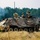 1-68 AR conducts an Abrams platoon live-fire exercise The image shows a wheeled armored personnel carrier (APC), likely an M1126 Stryker or similar 8-wheeled vehicle, draped with camouflage netting in an open field training environment. Three soldiers in camouflage uniforms are visible — two atop the vehicle and one on the ground beside it. Smoke is visible in the background, suggesting an active training exercise or field operation.