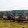 1-68 AR conducts an Abrams platoon live-fire exercise Three main battle tanks, identifiable as M1 Abrams variants, are positioned in a line across an open field with a treeline in the background. Multiple soldiers are visible on top of the tanks, appearing to conduct maintenance or preparation activities. The tanks are equipped with camouflage netting and appear to be in a field training or exercise environment.