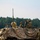1-68 AR conducts an Abrams platoon live-fire exercise The image shows military personnel positioned atop an armored vehicle draped with camouflage netting, likely during a training exercise or field operation. Two soldiers are visible in the vehicle's open hatch area, with one using binoculars or optical equipment. The setting appears to be a military training ground with a treeline and open field in the background.
