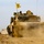 1-68 AR conducts an Abrams platoon live-fire exercise A U.S. Army M1 Abrams main battle tank is moving forward across a dusty, sandy terrain, kicking up a significant dust cloud from its tracks. A yellow unit guidon flag is mounted on the turret, and at least one crew member is visible in the commander's hatch. A second armored vehicle is partially visible in the background.