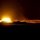 1-68 AR conducts an Abrams platoon live-fire exercise A nighttime image showing what appears to be a large muzzle flash or explosion from a military vehicle, likely a tank firing its main gun. The intense bright orange and white light illuminates the surrounding terrain and silhouettes of armored vehicles. Additional light sources are visible in the far right of the frame.