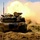 1-68 AR conducts an Abrams platoon live-fire exercise A main battle tank is captured mid-firing, with a large muzzle blast and fireball visible to the right side of the vehicle. The tank is traversing sandy, disturbed terrain while its main gun is being discharged. Dust and smoke surround the vehicle, indicating active combat training or live-fire exercise conditions.