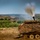 1-68 AR conducts an Abrams platoon live-fire exercise A military armored vehicle in the foreground is covered with camouflage netting and emitting smoke from its exhaust stack, suggesting active engine operation. In the background, at least two additional armored vehicles are visible on a rolling, semi-arid training terrain. The scene appears to be a military exercise or maneuver on an open training ground.