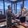 Two military officers are shaking hands in what appears to be a senior officer's office or command suite with large windows overlooking a harbor or waterfront area. One officer is wearing US Navy NWU Type III (digital camouflage) uniform, while the other is in a white naval dress uniform with rank insignia and medal ribbons consistent with a senior officer. The setting suggests a formal bilateral military courtesy visit or exchange.