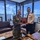 Two military officers, one in US Navy digital camouflage uniform and one in a white naval dress uniform, are jointly holding a commemorative wooden plaque in what appears to be a senior officer's office. An Australian flag is visible in the background, along with framed naval vessel artwork on the walls and a harbor/shipyard view through large windows. The setting suggests a formal bilateral military exchange or courtesy visit.