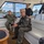 Several military personnel in various uniforms are seated and standing aboard a civilian motor yacht on a harbor waterway. The group appears to be on an official tour or transit, with a cityscape visible through the vessel's windows. At least four distinct military uniforms are visible, suggesting a multinational or multi-service gathering.