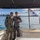 Two military personnel in different camouflage uniforms pose together on the stern of a motorboat on Sydney Harbour, Australia. One individual wears what appears to be a US Navy NWU Type III uniform, while the other wears an Australian Defence Force (AUSCAM) pattern uniform. The Australian White Ensign (Royal Australian Navy flag) is visible flying from the vessel, with the Sydney CBD skyline and Harbour Bridge visible in the background.