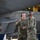 Three U.S. Air Force personnel in OCP (Operational Camouflage Pattern) uniforms are working in what appears to be an aircraft hangar. Two individuals in the foreground are engaged in conversation while wearing communication headsets, with one gesturing with his finger. A grey military aircraft, likely an F-22 Raptor, is visible in the background.