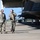 Two military personnel in camouflage uniforms are walking on an airfield tarmac beneath the wing of a military aircraft, appearing to conduct a pre-flight or maintenance inspection. Both individuals are wearing hearing protection headsets, with one gesturing as if explaining something to the other. Ground support equipment and a hangar structure are visible in the background.