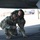 Two military personnel in US Army/Air Force OCP (Operational Camouflage Pattern) uniforms are crouching beneath an aircraft, conducting what appears to be a pre-flight or maintenance inspection. The foreground individual is wearing aviation headset/ear protection and holding a cable, while both are closely examining the underside or landing area of the aircraft. The setting appears to be a military airfield or flight line.