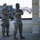 Two military personnel in OCP (Operational Camouflage Pattern) uniforms are standing on what appears to be a flight line or aircraft ramp area. Both individuals have their arms crossed and are wearing aviation headsets, suggesting they are monitoring aircraft operations. The setting appears to be a military air base during daylight hours.