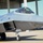 A fifth-generation stealth fighter aircraft, identifiable as an F-22 Raptor, is taxiing on a military airfield apron. A pilot in full flight gear including helmet and oxygen mask is visible in the cockpit. A large aircraft hangar is visible in the background.