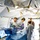 Military medical personnel are performing a surgical or medical procedure inside a field hospital tent. Three individuals in surgical gowns and purple gloves work around a patient on a table draped in blue surgical draping, while two uniformed military observers stand nearby. The setting is a portable military medical facility with fluorescent lighting and white tent walls.