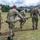 Military personnel in U.S. Navy and Marine Corps uniforms are conducting a medical casualty evacuation drill in an outdoor field setting. Three service members are carrying a simulated casualty on a litter/stretcher toward what appears to be a field medical station with camouflage netting and tents in the background. A fourth individual in a green t-shirt and Marine Corps trousers observes or assists nearby.